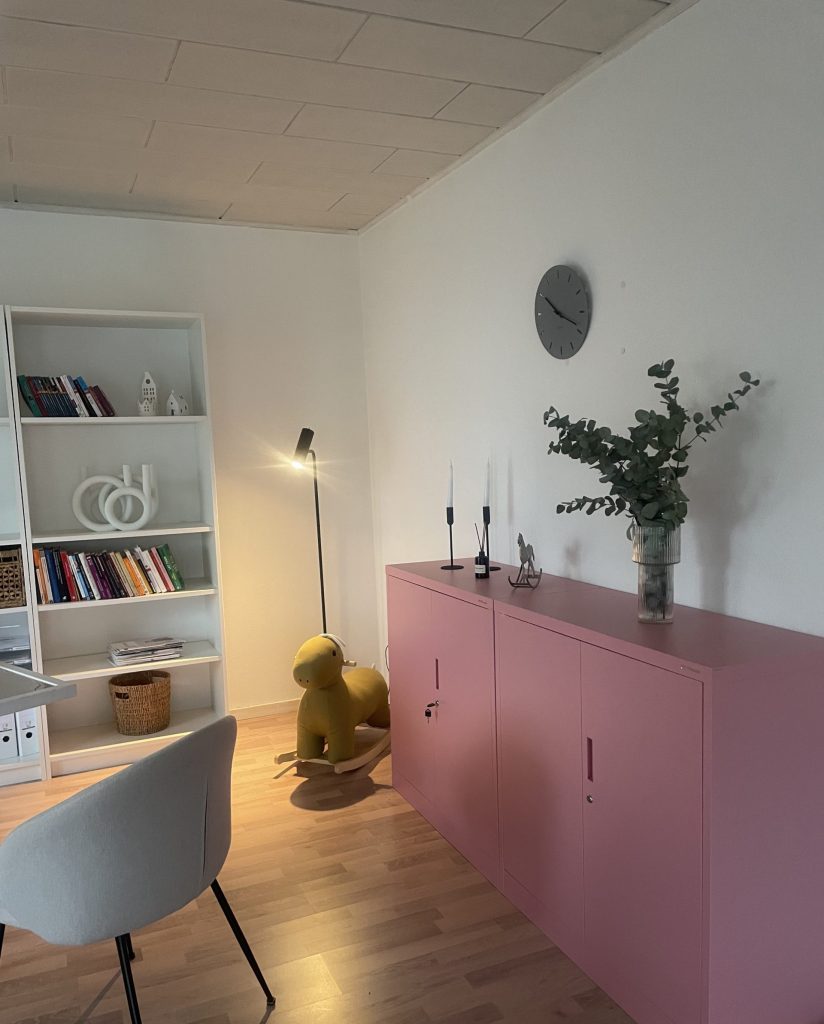 Ein Bild mit Regal, Uhr, Schaukeltier und Stuhl aus dem 1. Behandlungszimmer